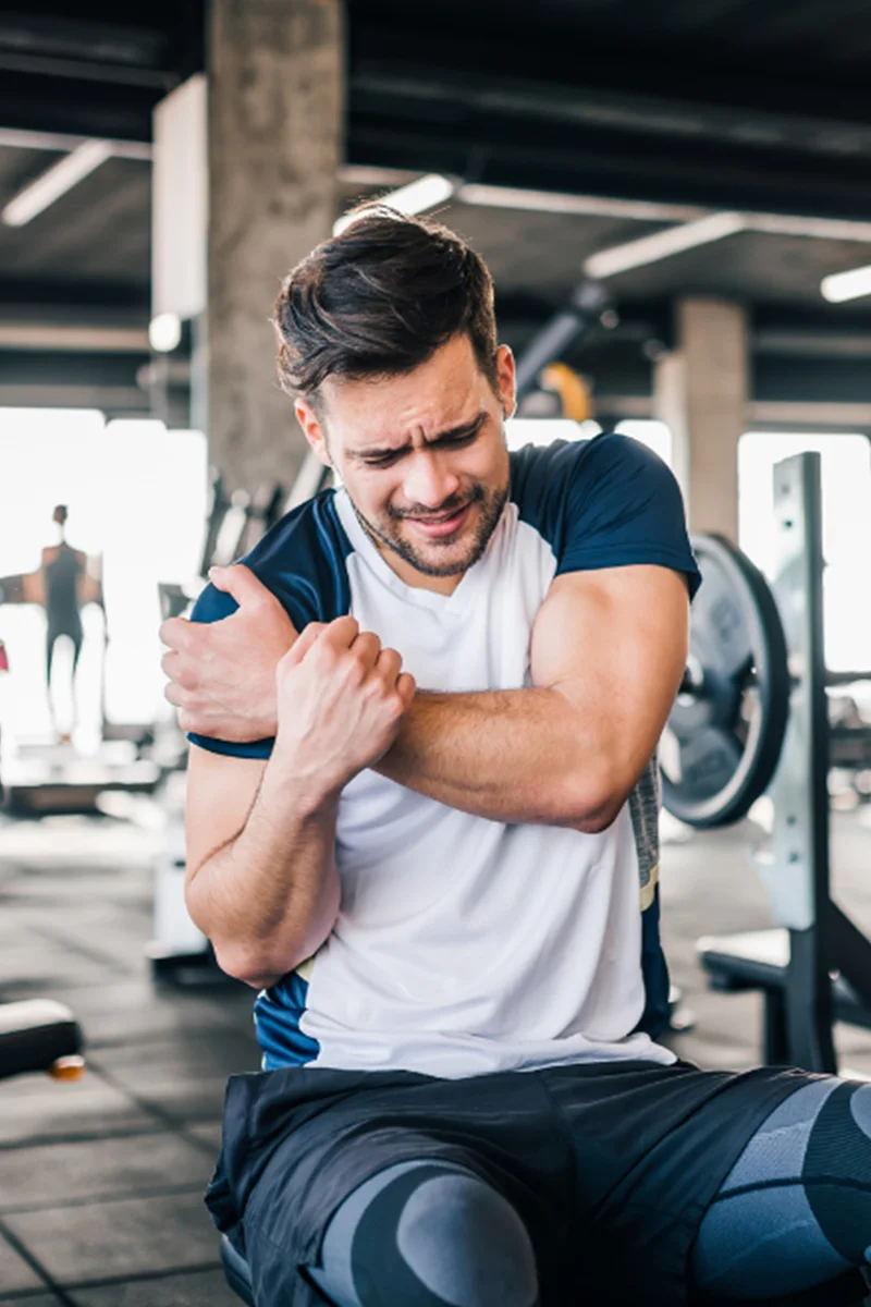 Váll- és könyökfájdalom sport közben: egy férfi edzőteremben fájlalja a vállát és a könyökét, teniszkönyök vagy vállprobléma miatt.